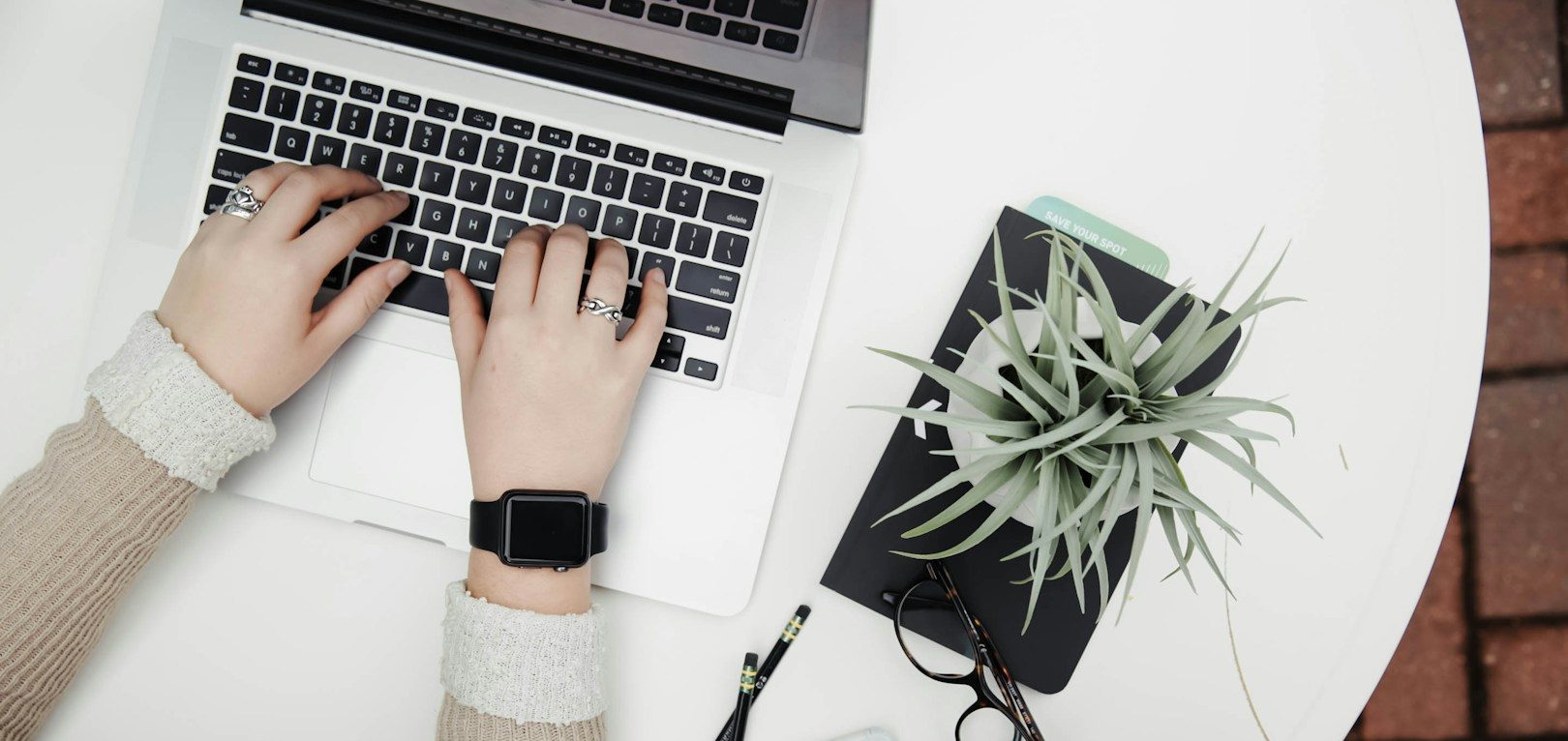 Mains sur un ordinateur portable, plante succulente et carnet sur une table blanche.