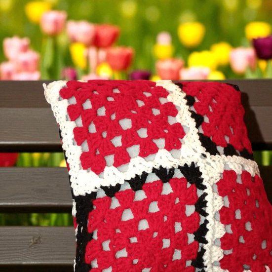 Coussin en crochet à motifs rouges, noirs et gris, posée sur un banc en bois.