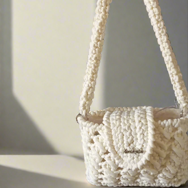Petit sac à main blanc en tricot, posé sur une surface avec une ombre légère. FeniCrochet