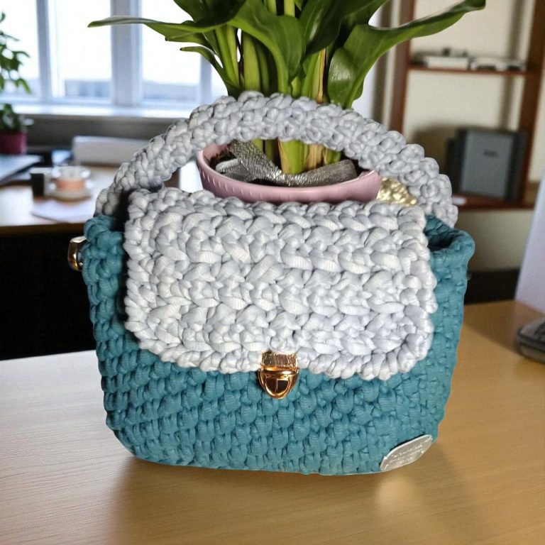Sac à main crocheté en bleu et gris avec une poignée beige, posé sur une table. Fenicrochet